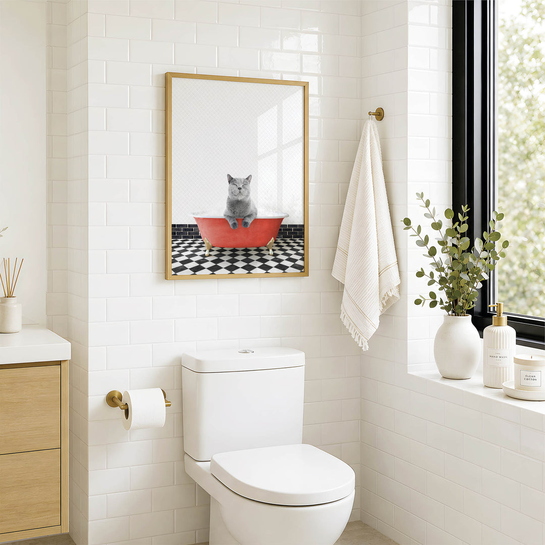 A bathroom with a white toilet, a framed picture of a cat sitting in a red bathtub, and a window with greenery on the sill.