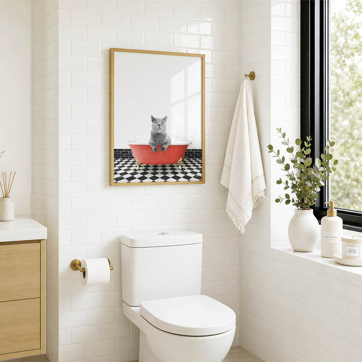 A bathroom with a white toilet, a framed picture of a cat sitting in a red bathtub, and a window with greenery on the sill.