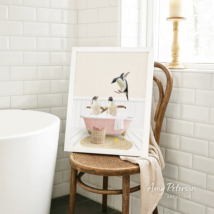 A framed image of two penguins sitting in a pink bathtub, surrounded by a basket of bath toys, is displayed on a wooden chair in a bathroom setting.