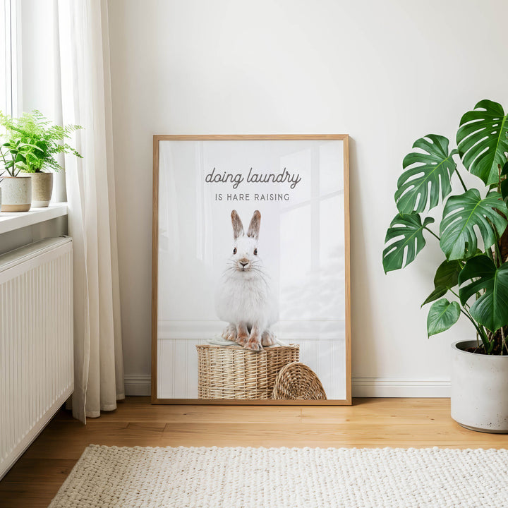 a framed poster of a white rabbit sitting in a wicker basket, placed on a wooden floor next to a potted plant.