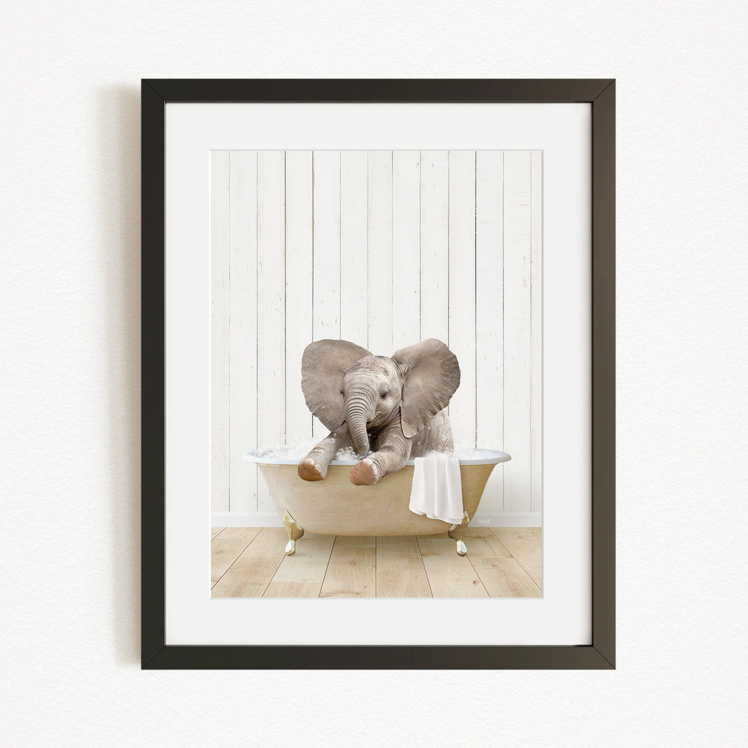 A baby elephant sitting in a bathtub, with its trunk raised and ears spread out, against a white wooden wall.