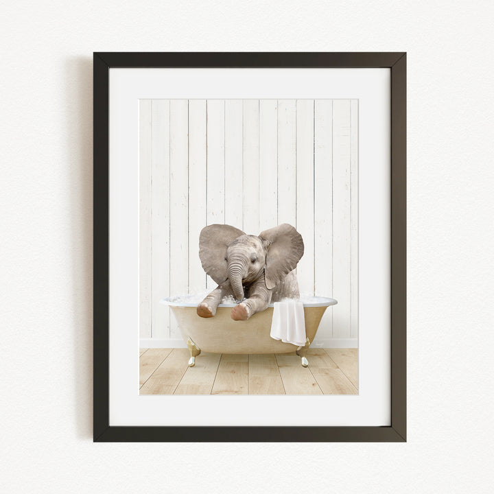 A baby elephant sitting in a bathtub, with its trunk raised and ears spread out, against a white wooden wall.