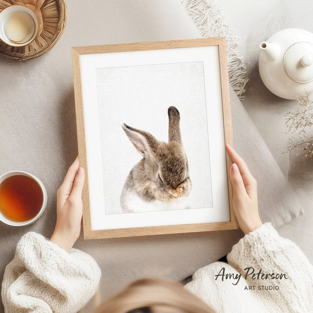 A person is holding a framed picture of a rabbit, with a teapot, a cup of tea, and a bowl of tea visible in the background.