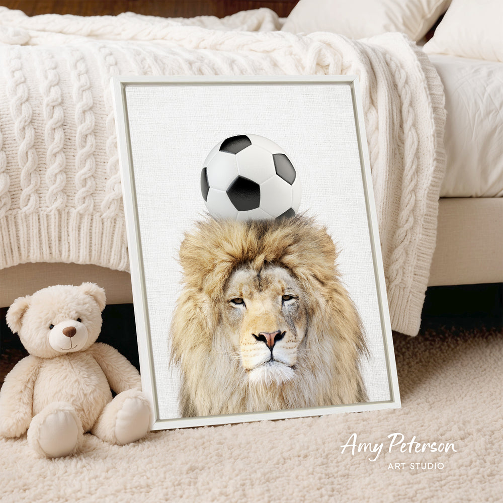 A framed print of a lion with a soccer ball on its head is displayed on a bed, accompanied by a teddy bear.
