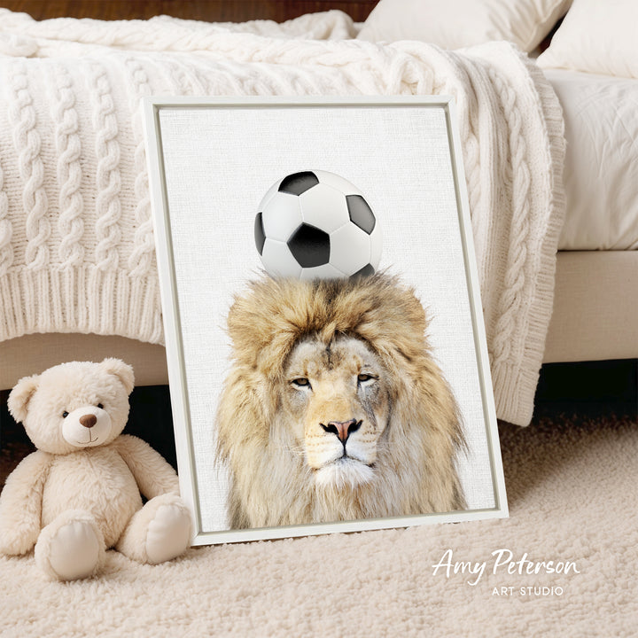A framed print of a lion with a soccer ball on its head is displayed on a bed, accompanied by a teddy bear.