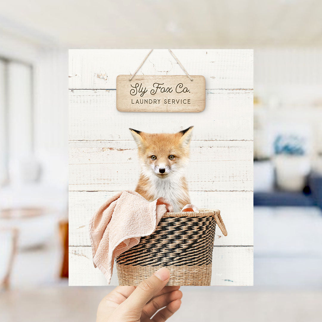 A hand holds up a framed image of a small orange fox sitting in a woven basket, with a sign above it that reads "Laundry Service".