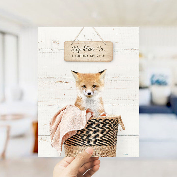 A hand holds up a framed image of a small orange fox sitting in a woven basket, with a sign above it that reads "Laundry Service".