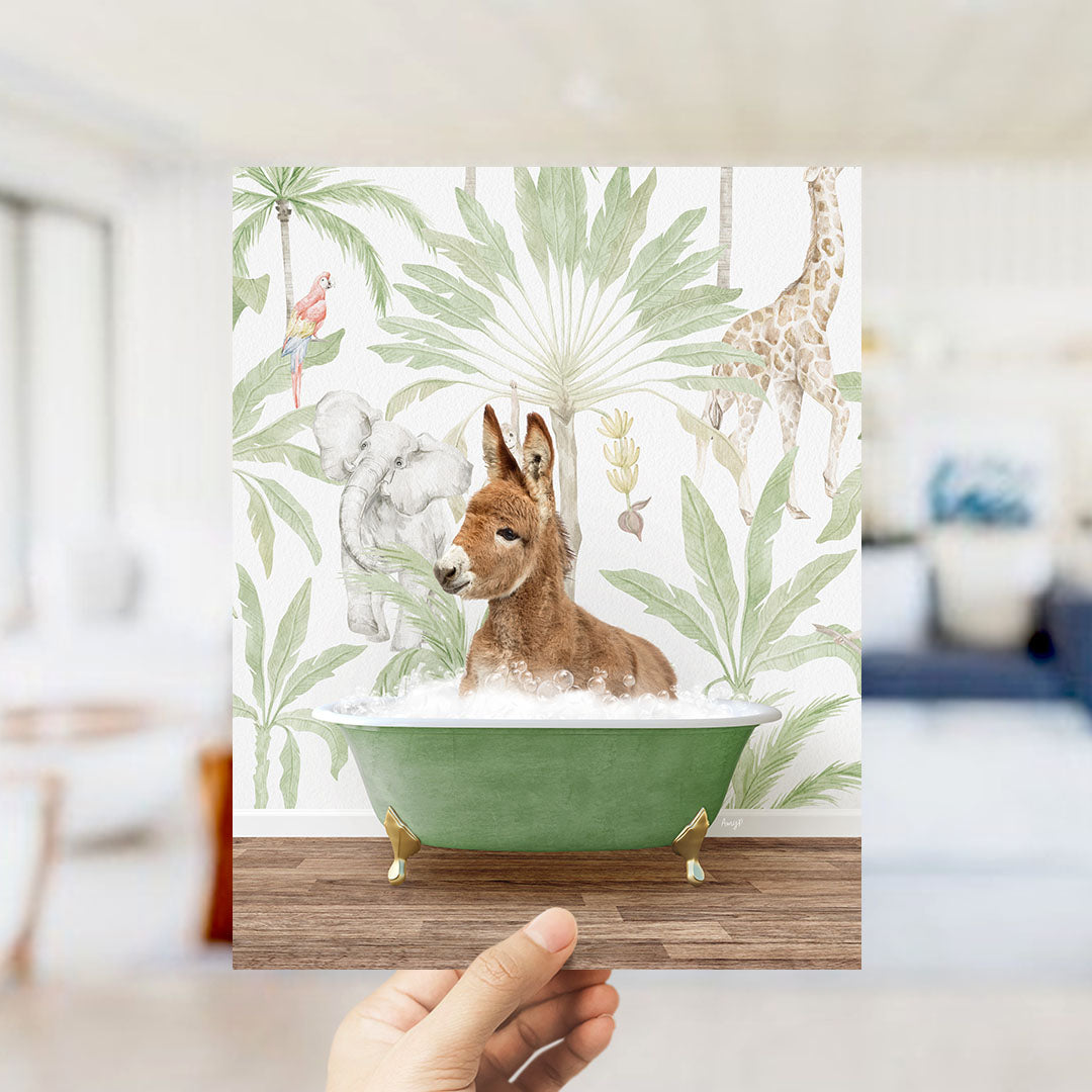 A hand holds up a framed print featuring a whimsical scene of a donkey sitting in a bathtub surrounded by tropical foliage, including a giraffe and palm trees.