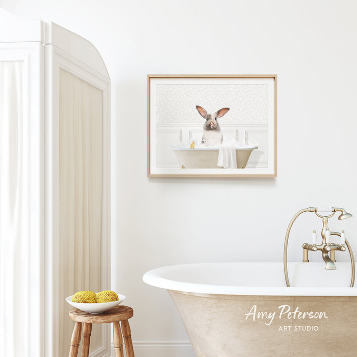 A framed image of a rabbit sitting in a bathtub is hanging on a white wall above a freestanding bathtub in a bathroom.