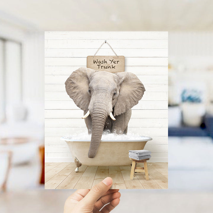 A hand holds up a framed image of an elephant sitting in a bathtub, with a sign above it that says "Wash Yer Trunk".