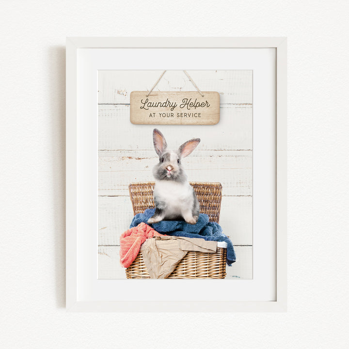 A white rabbit sitting in a wicker basket filled with clothes, with a sign above that reads "Laundry Helper at your service".