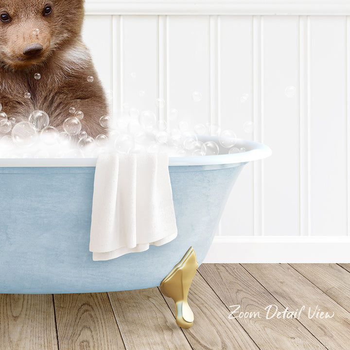 A brown bear sitting in a blue bathtub filled with bubbles, with a towel and gold bathtub leg visible.