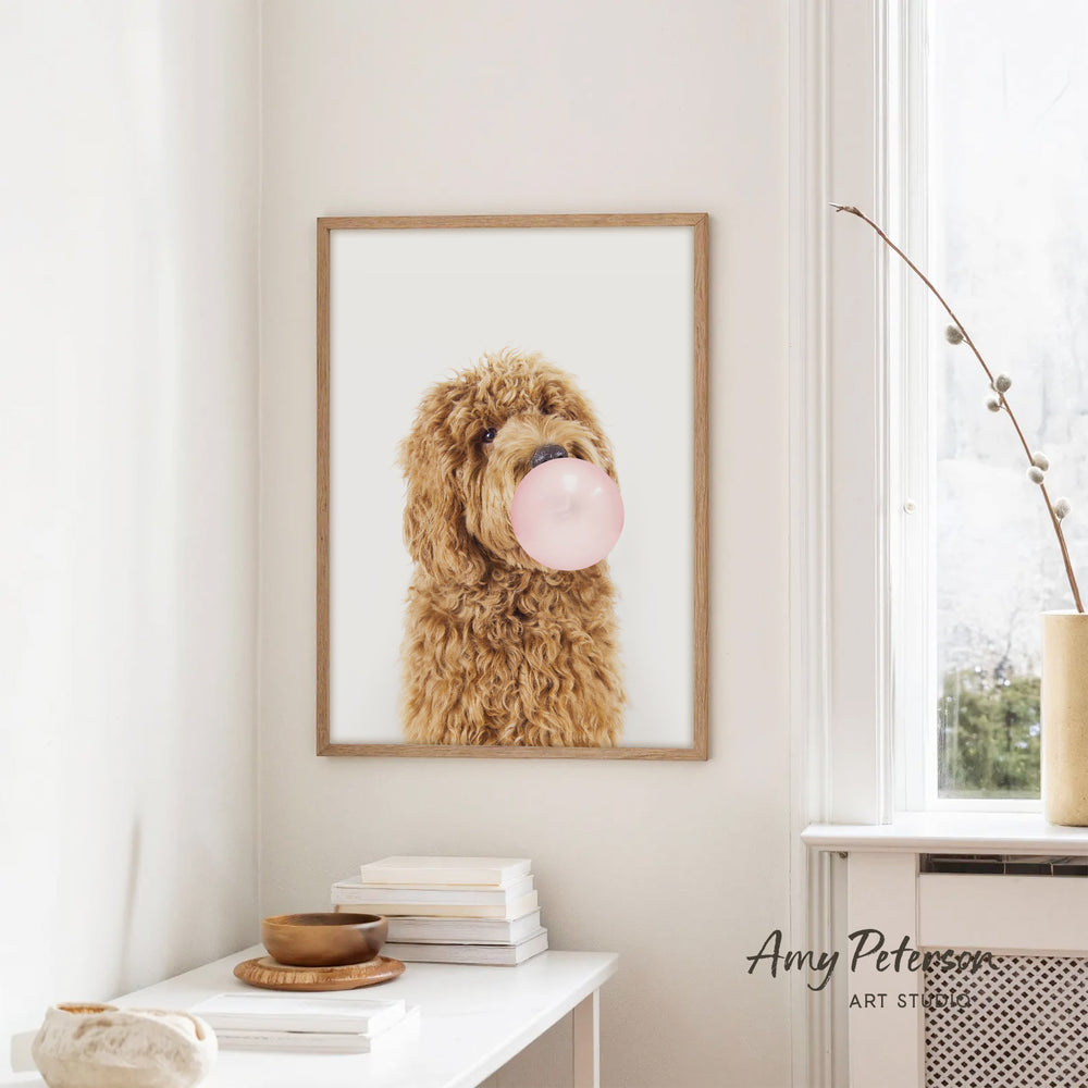 A framed image of a brown dog blowing a bubble gum bubble is hanging on a white wall in a room with a white table and a window.