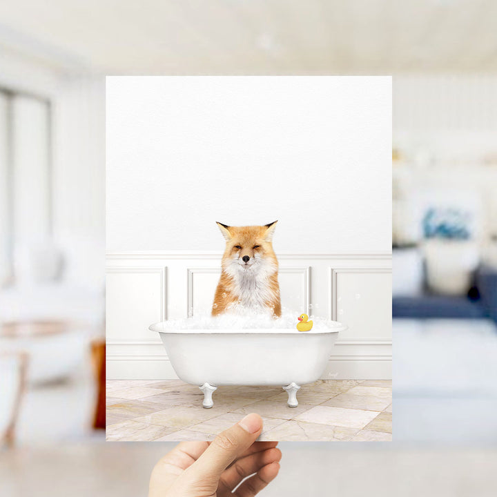 A hand holds up a framed image of a fox sitting in a bathtub filled with bubbles, with a rubber duck floating nearby.