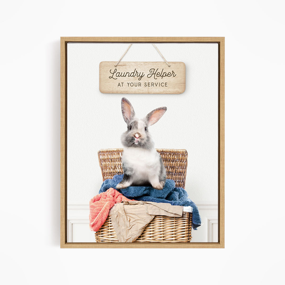 A gray and white rabbit sitting in a wicker laundry basket filled with clothes, with a wooden sign above that reads "Laundry Helper at your service".