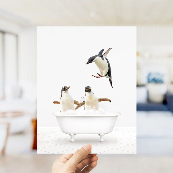 A hand holds up a framed image depicting two penguins sitting in a bathtub, with one penguin flying out of the tub.