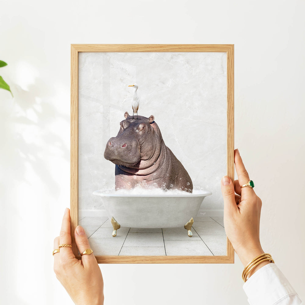 A person's hands are holding a framed picture of a hippopotamus sitting in a bathtub, with a bird perched on its head.