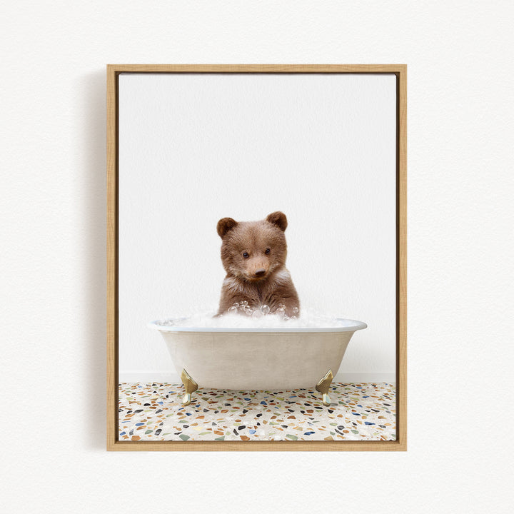 A cute brown bear cub sitting in a white bathtub filled with water, looking directly at the camera.