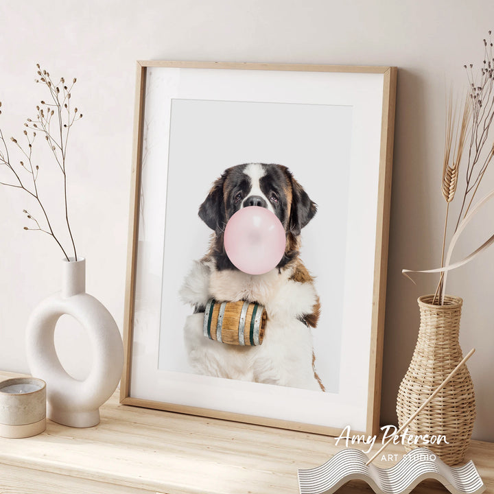 A framed photograph of a dog blowing a bubble with a wooden barrel around its neck, sitting on a wooden surface next to a white vase with dried flowers.
