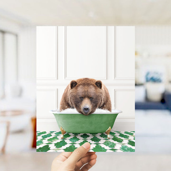 A hand holds up a framed image of a brown bear sitting in a green bathtub filled with bubbles, with a white wall and green and white patterned floor in the background.