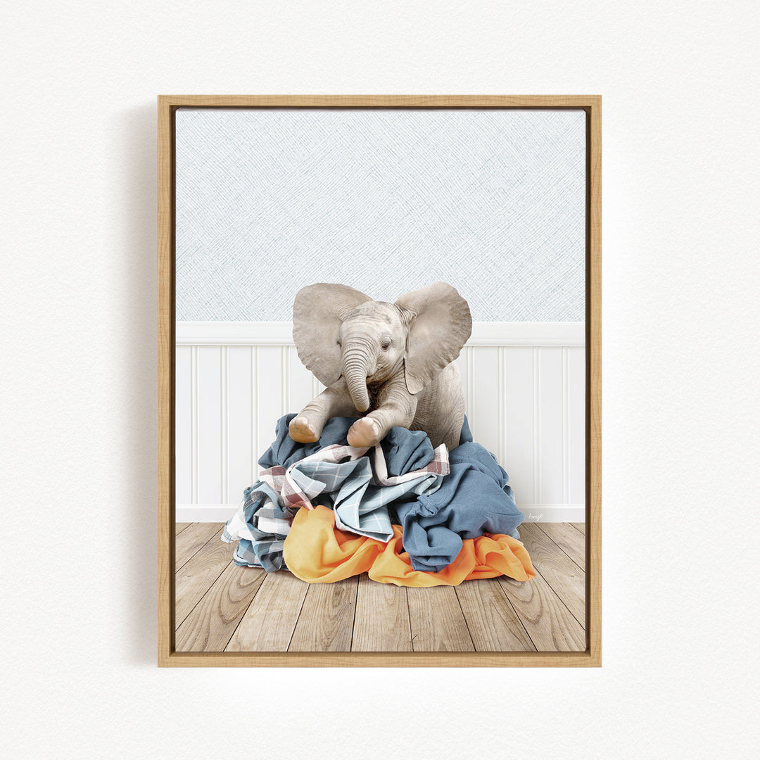 A stuffed elephant sits atop a pile of clothes on a wooden floor, with a white wall and a wooden panel in the background.