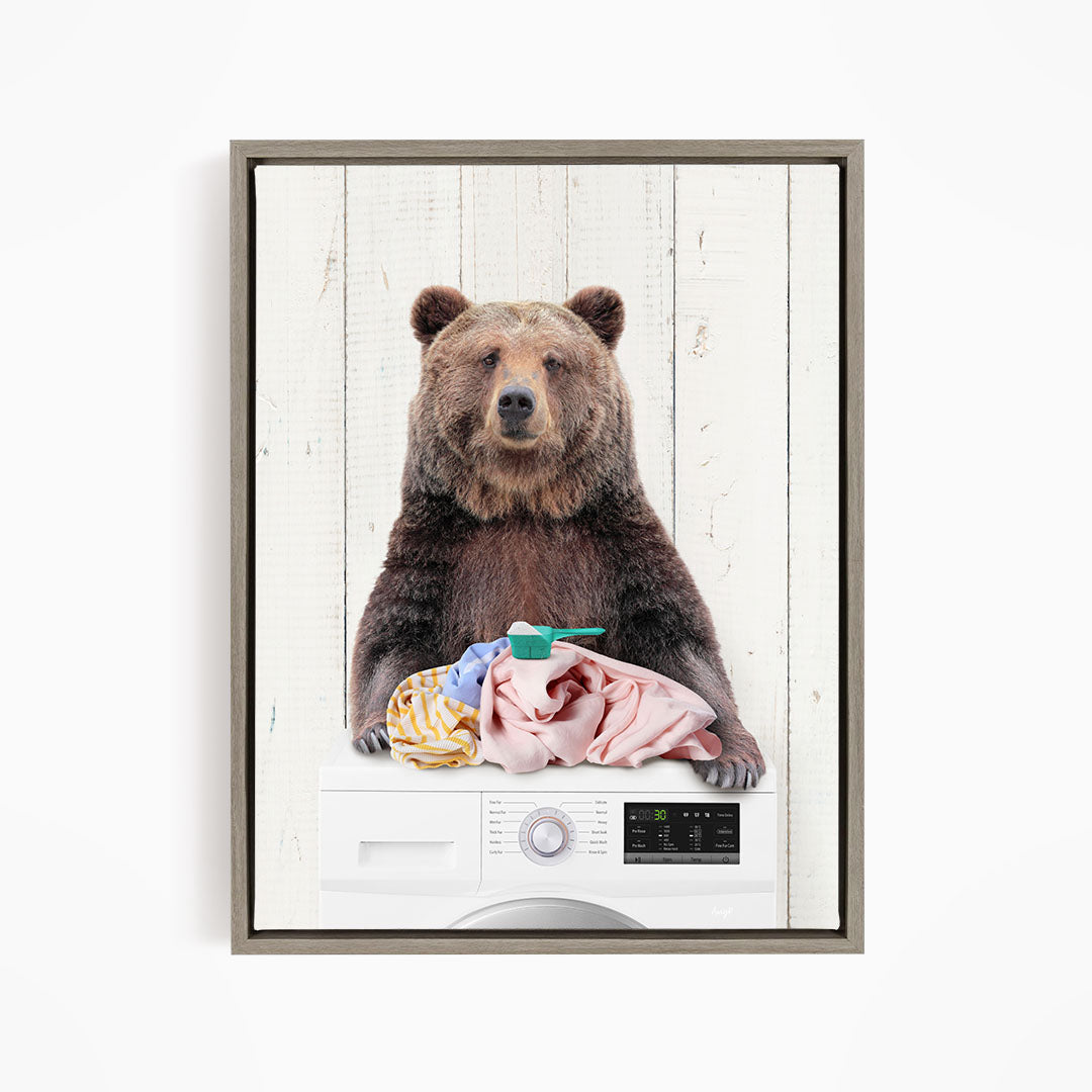 A brown bear sitting on top of a washing machine, holding a pink towel and a green object, with a white wooden background.