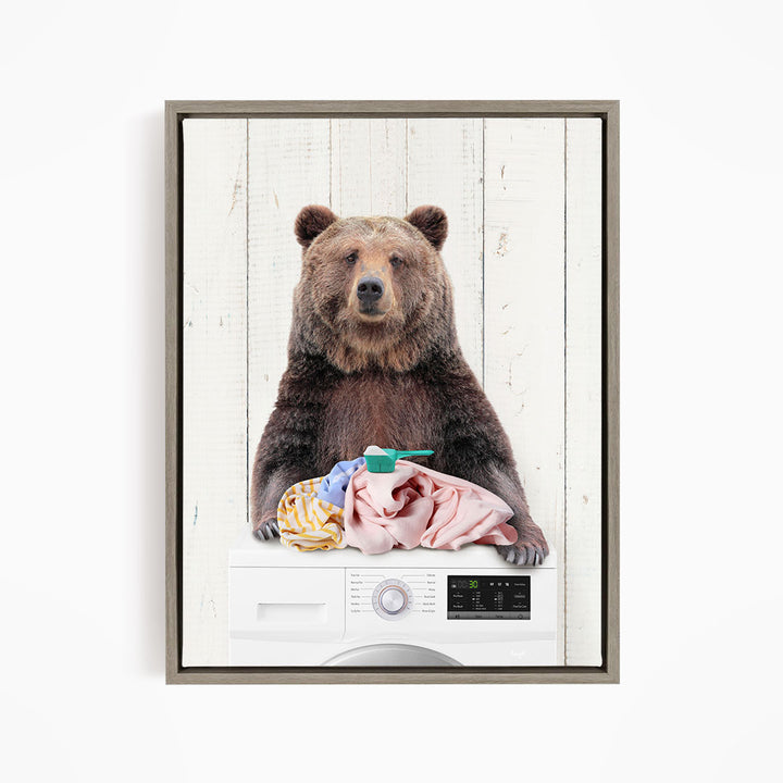A brown bear sitting on top of a washing machine, holding a pink towel and a green object, with a white wooden background.