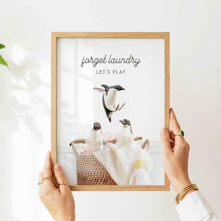 A person's hands are holding a picture frame with a photograph of two penguins sitting in a basket, with the text "forget laundry let's play" written above the image.