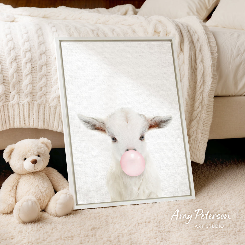 A framed image of a white goat blowing a bubble with a pink bubble gum bubble is displayed on a white bedspread, accompanied by a teddy bear.