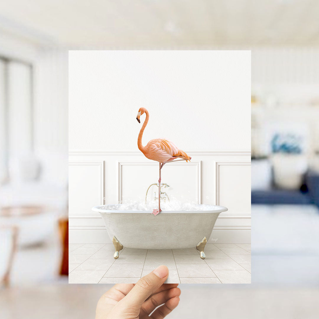 A hand holds up a framed image of a flamingo standing on the edge of a bathtub filled with water.