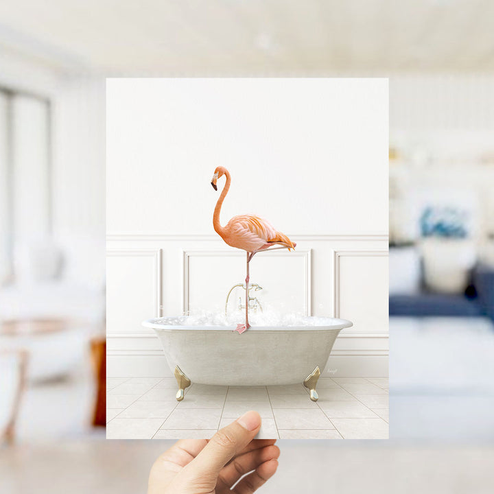 A hand holds up a framed image of a flamingo standing on the edge of a bathtub filled with water.