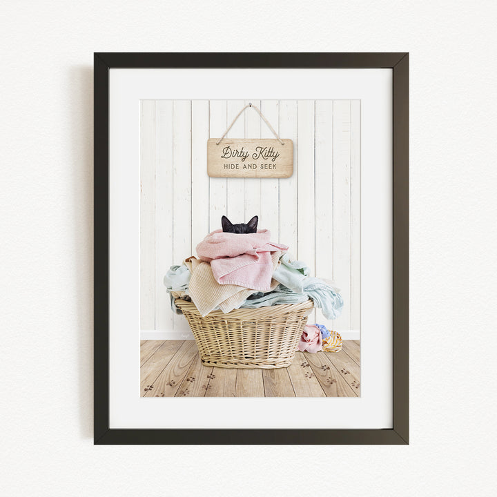 A framed photograph of a cat sitting in a basket filled with laundry, with a sign above the basket that reads "Dirty Kitty HIDE AND SEEE".