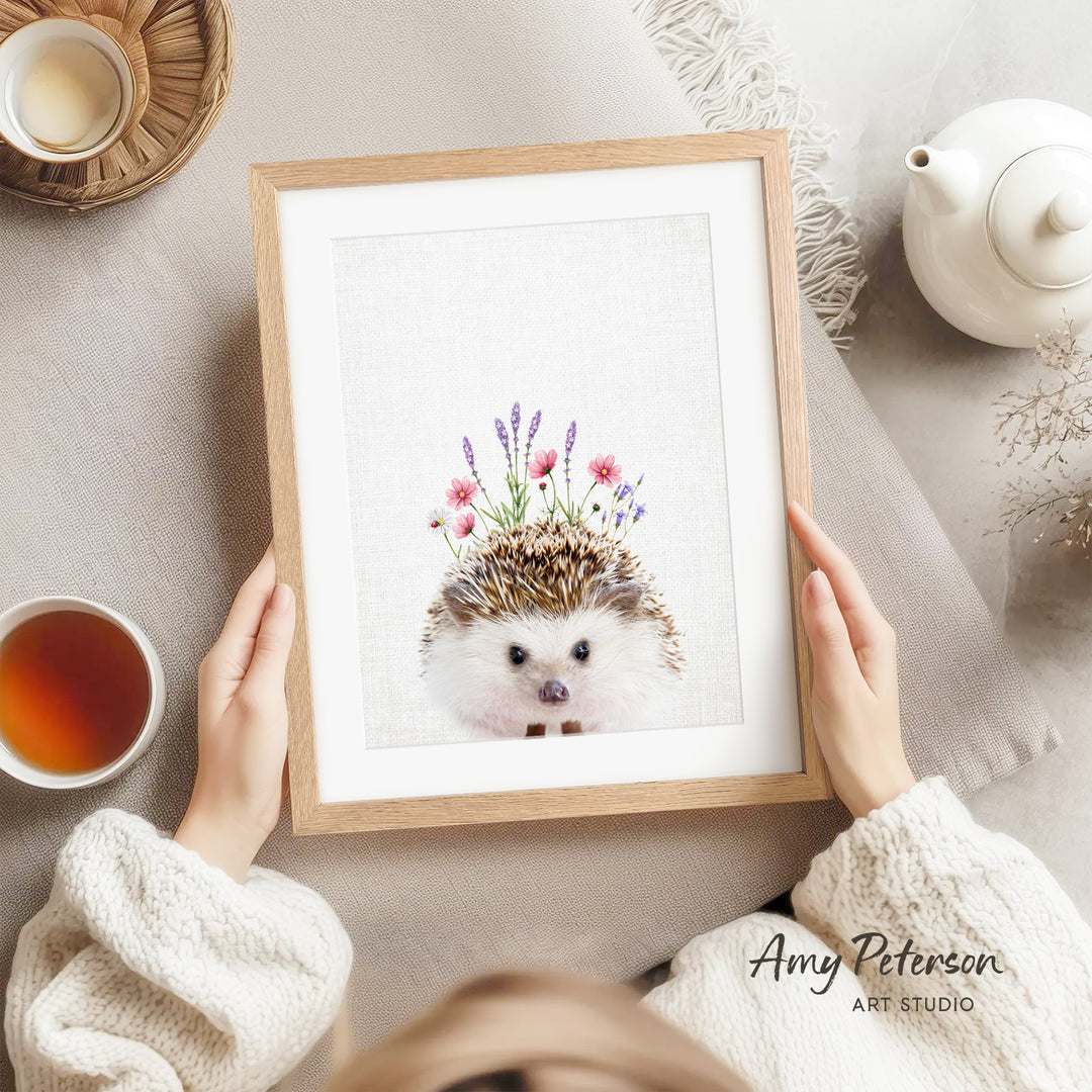 A person is holding a framed print of a hedgehog with flowers on its head, surrounded by various objects such as a teapot, a cup, and a bowl.