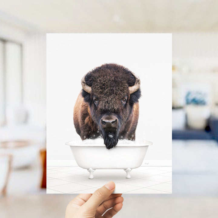 A hand holds up a framed image of a bison with a long beard, sitting in a white bathtub.