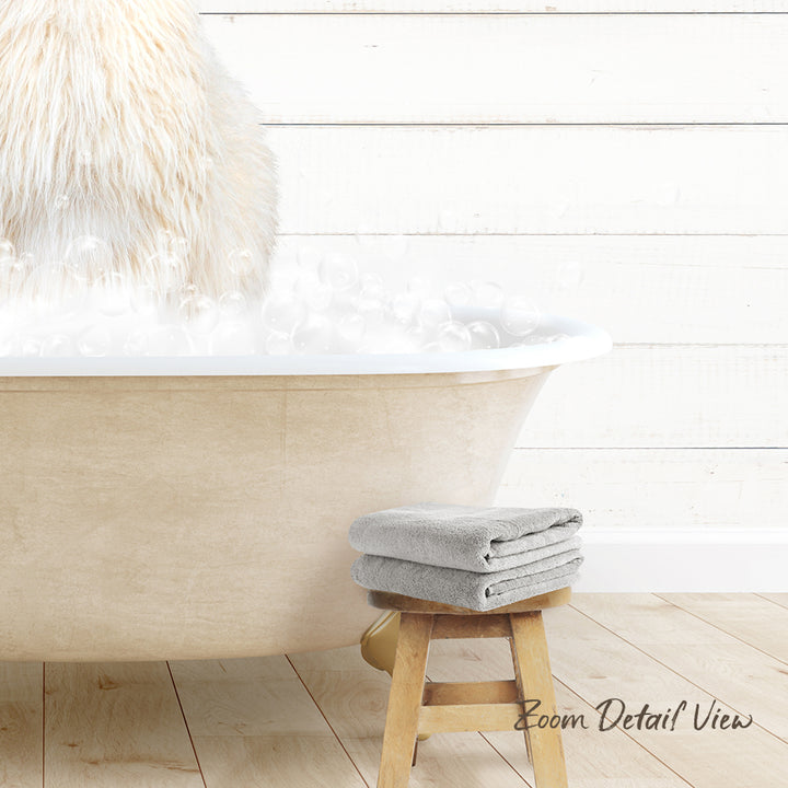 A fluffy white dog is sitting in a bathtub filled with bubbles, with a wooden stool and towels nearby.