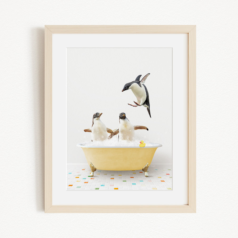 A framed image depicting two penguins sitting in a yellow bathtub, with one penguin standing on its hind legs and another penguin sitting on its belly, while a third penguin jumps into the tub.
