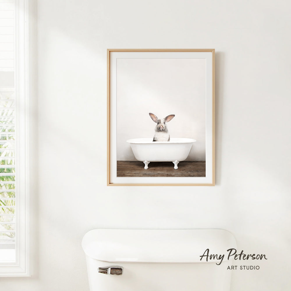 A framed image of a rabbit sitting in a white bathtub is hanging on a white wall.