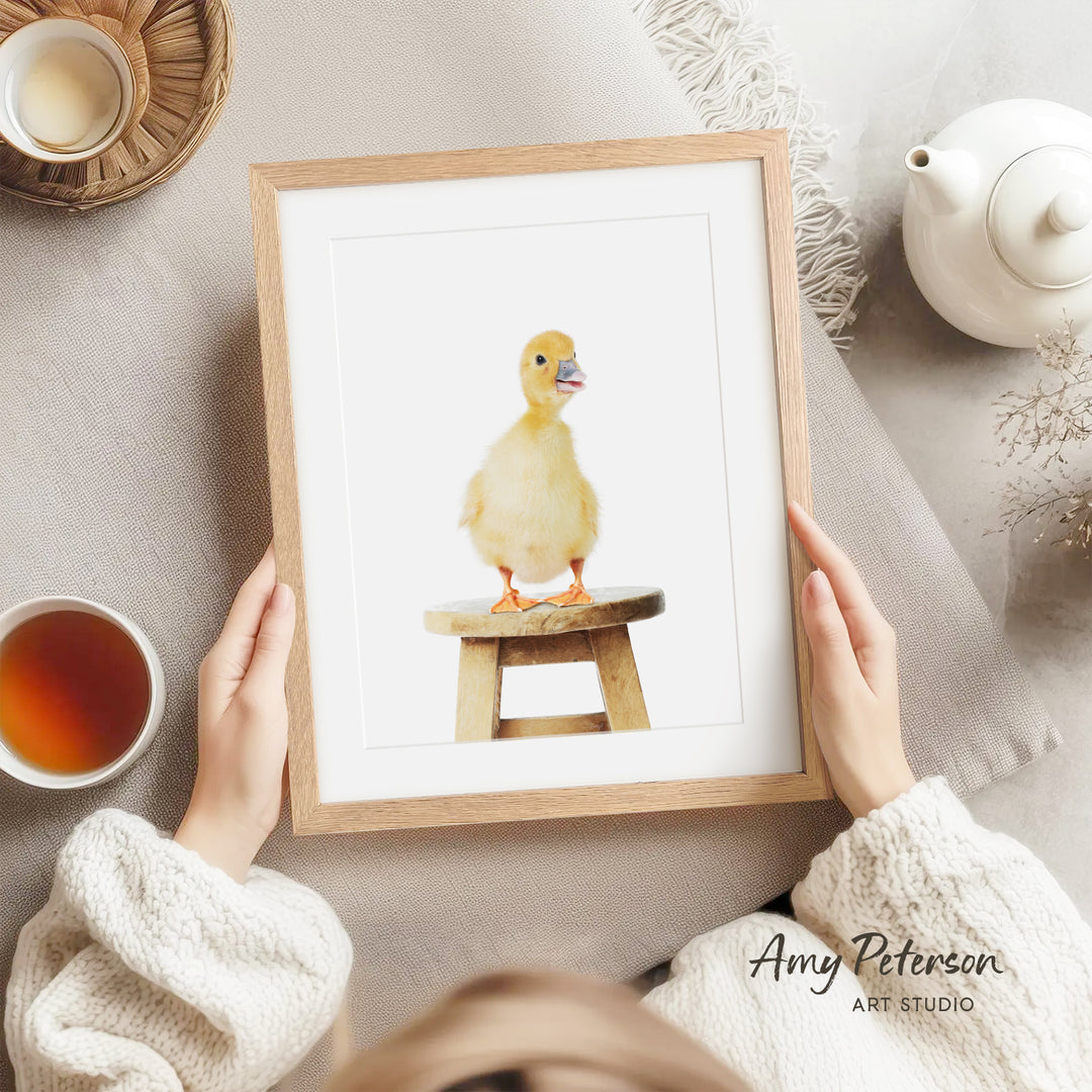 A person is holding a framed picture of a yellow duck sitting on a wooden stool, with a teapot, a cup of tea, and a bowl of tea nearby.