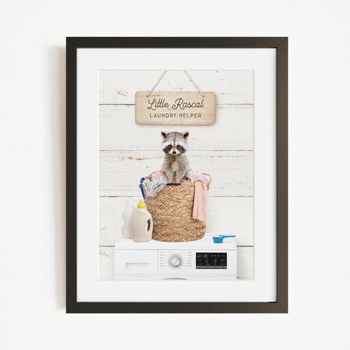 A framed image depicting a raccoon sitting in a woven laundry basket, surrounded by various household items such as a washing machine and a bottle of detergent.