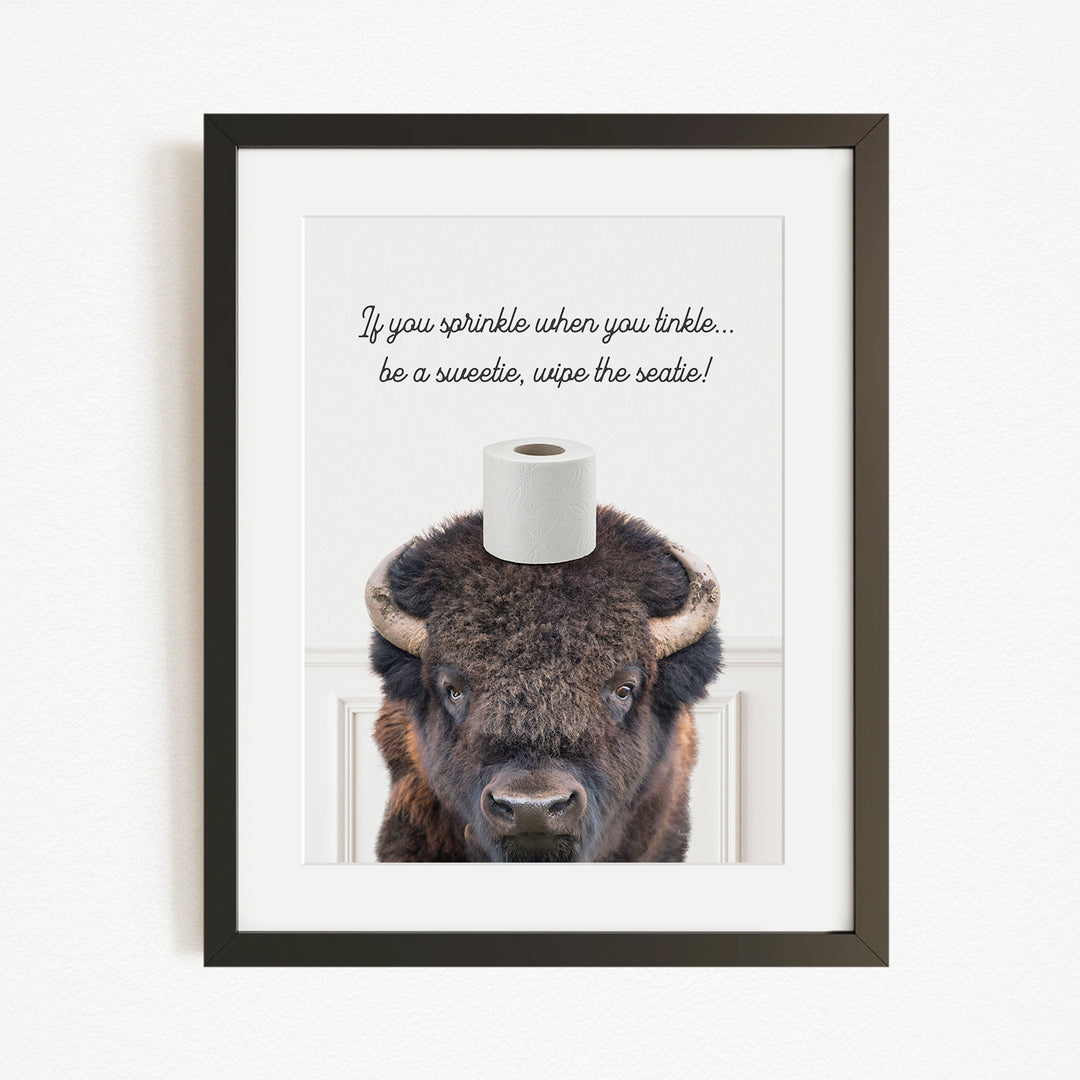 A framed image of a bison with a roll of toilet paper on its head, accompanied by the text "If you sprinkle when you tinkle... be a sweetie, wipe the seattle!"