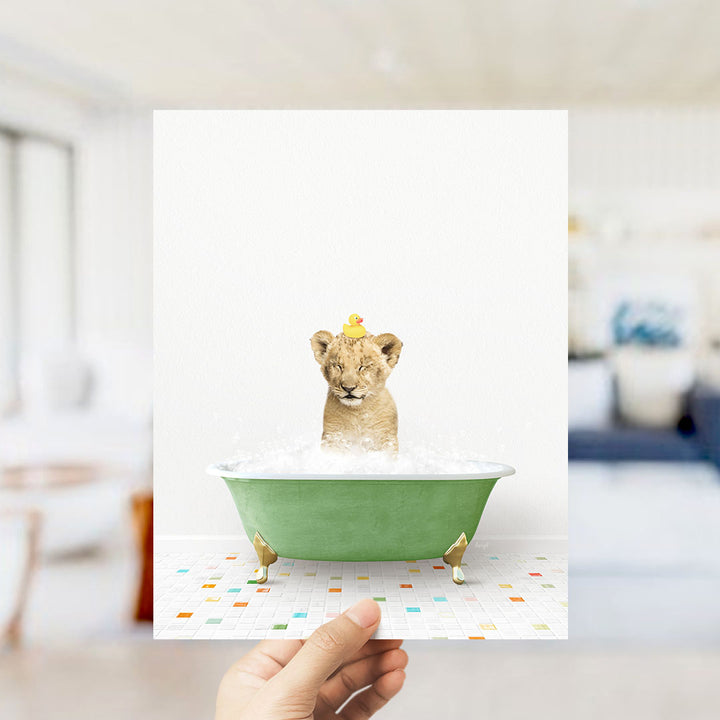 A hand holds up a framed image of a lion cub sitting in a green bathtub filled with bubbles, with a yellow rubber duck perched on its head.