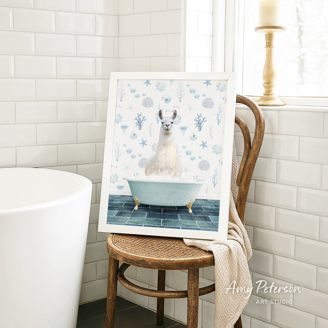 A framed image of a white llama sitting in a blue bathtub, surrounded by blue and white floral patterns, is displayed on a wooden chair in a bathroom setting.