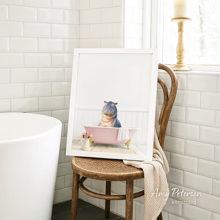 A framed image of a hippopotamus sitting in a pink bathtub, with a towel draped over the back of a wooden chair in a bathroom setting.