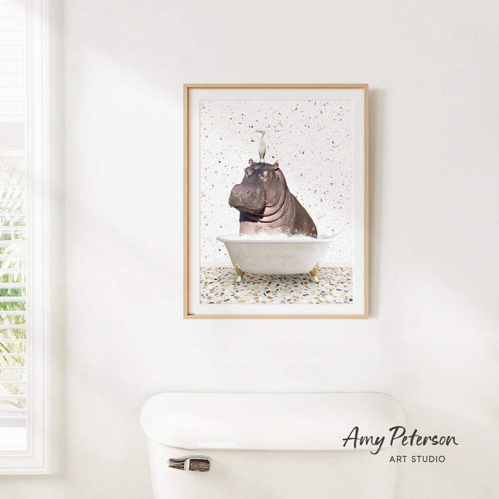 A framed print of a hippopotamus sitting in a bathtub is hanging on a white wall.