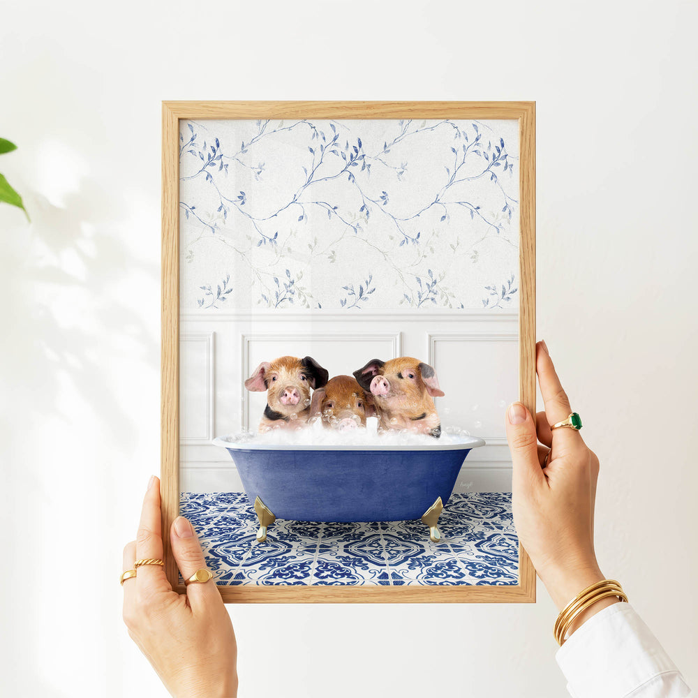 A person's hands are holding a framed picture of three adorable pigs sitting in a blue bathtub, with a floral pattern on the wall behind them.