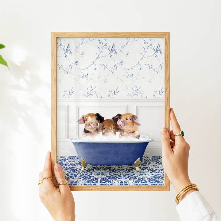 A person's hands are holding a framed picture of three adorable pigs sitting in a blue bathtub, with a floral pattern on the wall behind them.