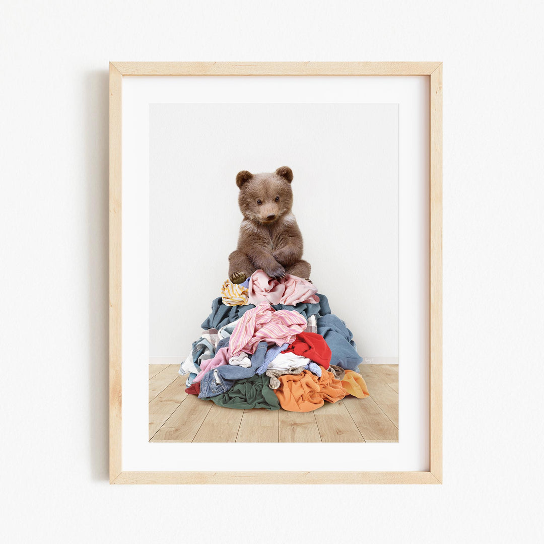 A brown bear sits atop a pile of colorful clothes on a wooden floor.