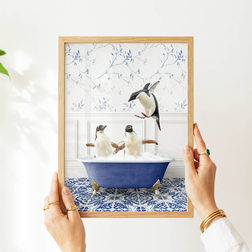 A person's hands are holding a framed picture of two penguins sitting in a blue bathtub, with one penguin flying towards the other.