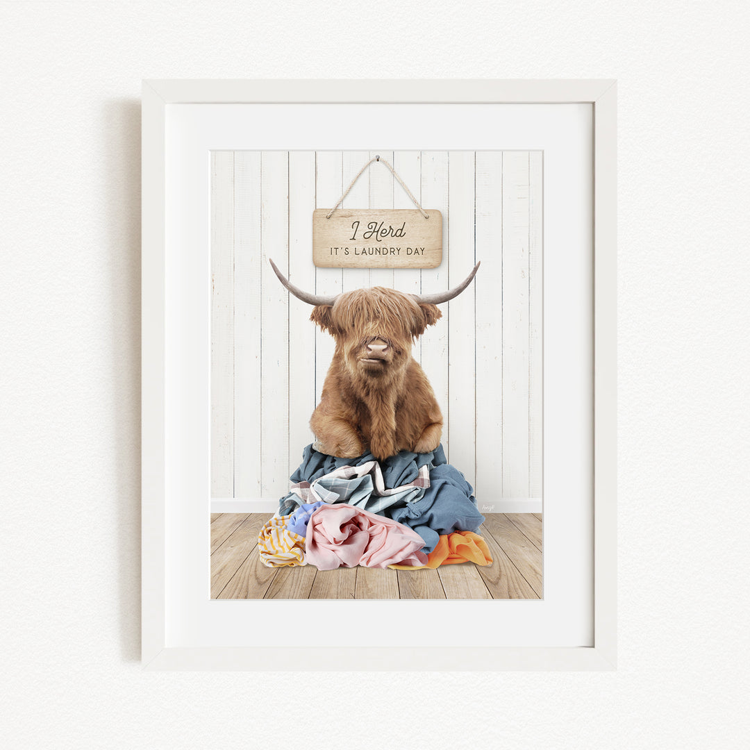 A framed image of a Highland cow with large horns sitting on a pile of clothes, with a sign above it that says "It's laundry day".