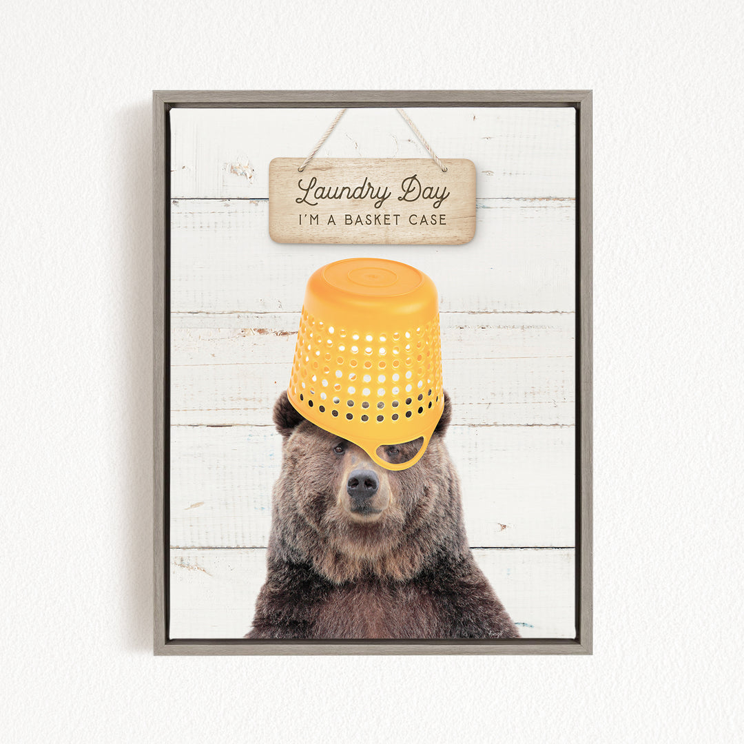 A framed image of a bear wearing a yellow plastic basket on its head, with a wooden sign above it that says "Laundry Day I'm a basket case".